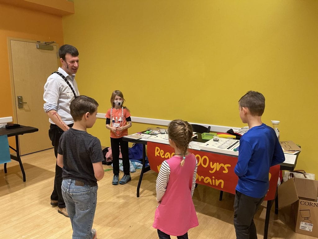 Dr. William Buscher, Assistant Professor in the Department of Genetics, talk with his daughter (in pink, left) and other children about neurodegeneration using a game built by Dr. Tom Mahan (postdoc in the Developmental Biology Department). Dr. Buscher sports a healthy neuron on his cheek and a degenerated neuron on his arm made with face paint.
